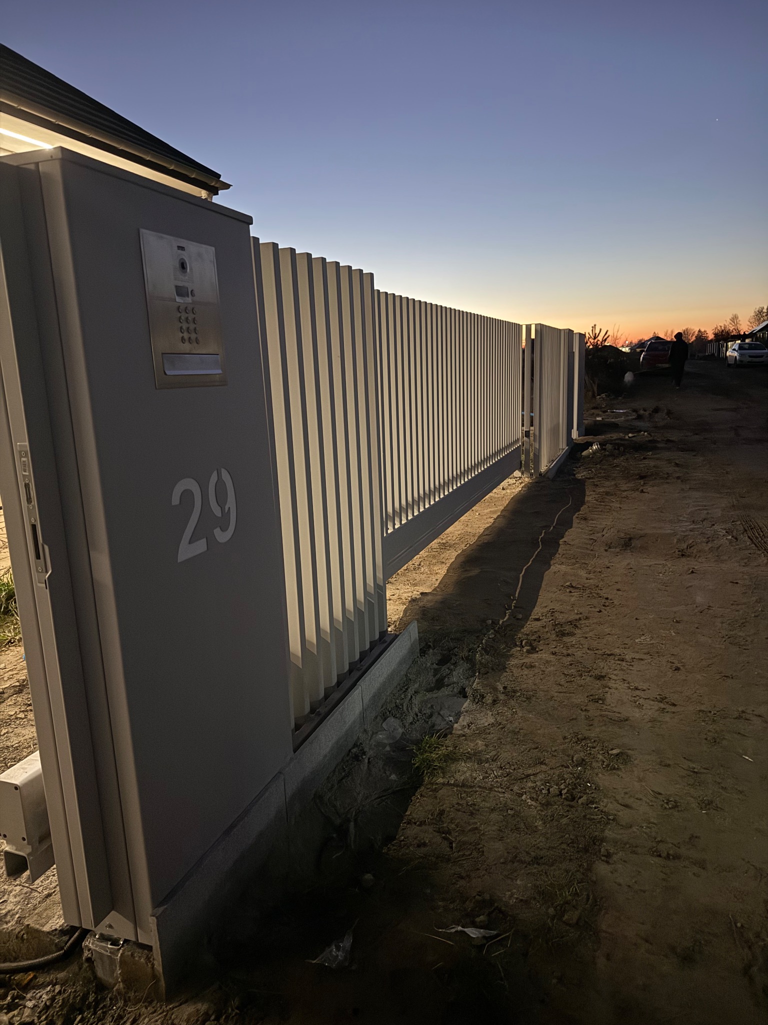 Nowoczesna, przesuwna brama wjazdowa z numerem domu '29' i domofonem, widziana o zmierzchu. Pionowe panele bramy w kolorze jasnoszarym.