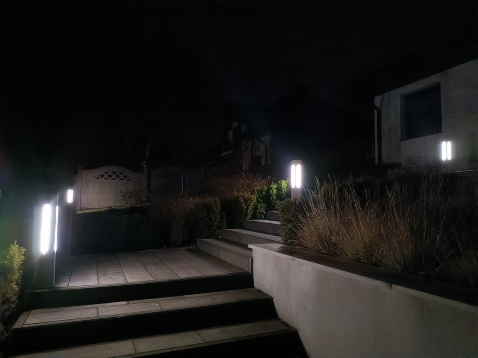 Oświetlona ścieżka z kamiennymi schodami prowadząca do domu nocą. Lampy ogrodowe po bokach ścieżki. Minimalistyczny design.