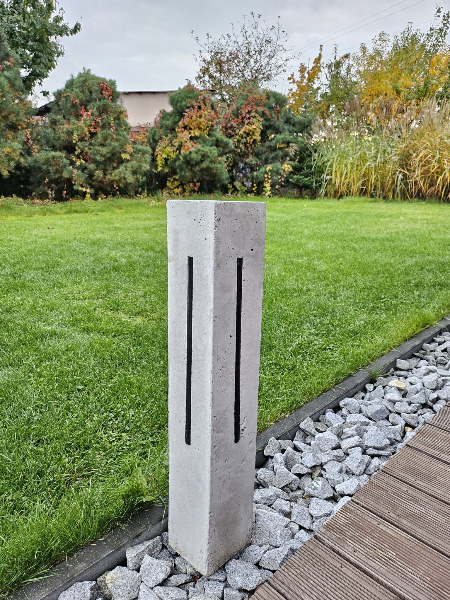 Betonowa lampa ogrodowa z dwoma szczelinami na światło, ustawiona na trawniku obok ścieżki z kamieni i drewnianego podestu, w tle zieleń drzew i krzewów.