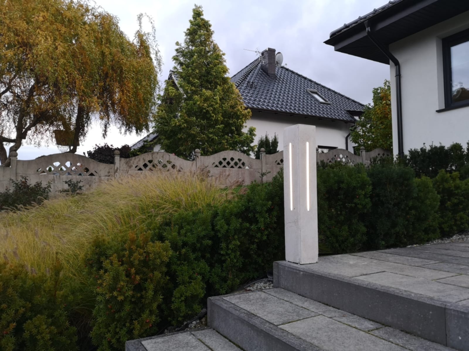 Nowoczesna lampa ogrodowa z betonu architektonicznego, oświetlająca schody w Ostrzeszowie. Minimalistyczny design, subtelne światło, tło z zieleni i domu.