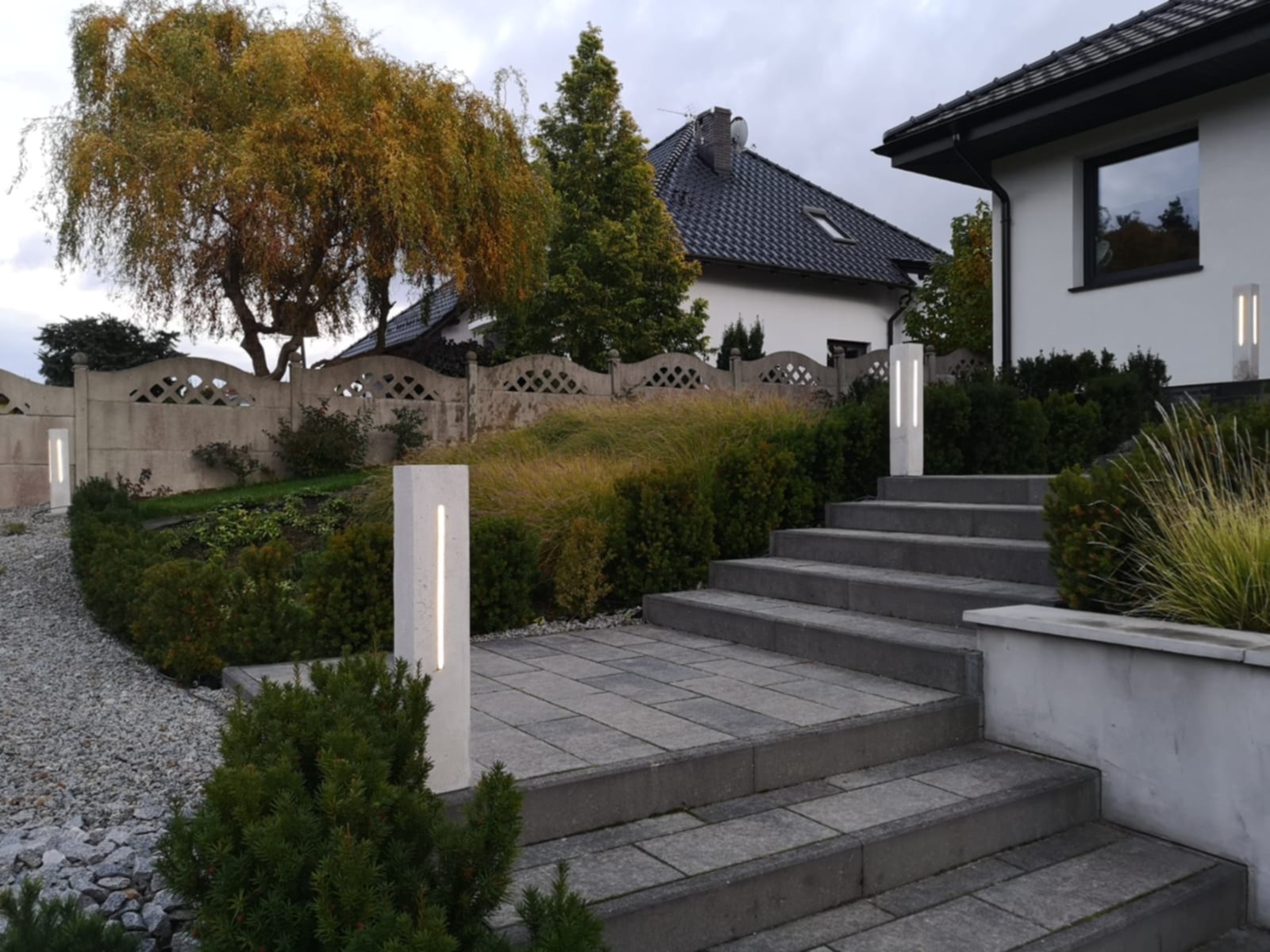 Oświetlenie ścieżki i schodów prowadzących do domu, z wykorzystaniem nowoczesnych, minimalistycznych lamp w kształcie słupków. Ogród z dekoracyjnymi roślinami.