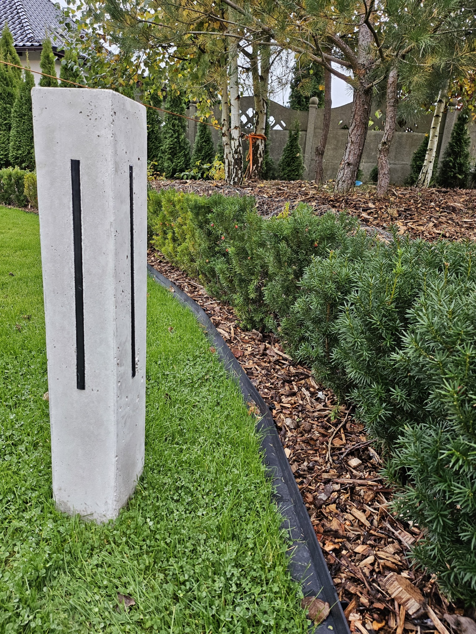 Betonowa lampa ogrodowa z czarnymi wstawkami na trawniku, obok rabaty z korą i iglakami. W tle drzewa i ogrodzenie.