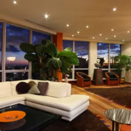 Elegancki salon z białym narożnikiem, dużymi oknami z widokiem na miasto o zmierzchu, egzotycznymi roślinami, drewnianą podłogą i dekoracyjnymi dodatkami, oświetlony punktowo.