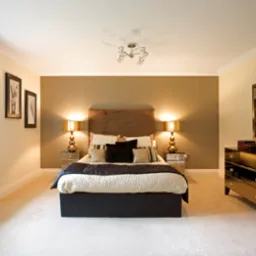 Elegancka sypialnia z łóżkiem małżeńskim, szafkami nocnymi z lampkami, komodą z lustrem i dekoracjami, utrzymana w tonacji beżu i brązu, z oknem z roletami rzymskimi.