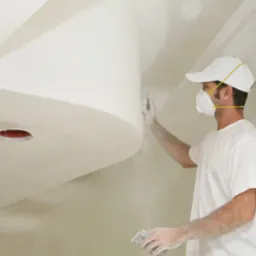 Pracownik w masce i czapce szlifuje gładź na suficie z elementami dekoracyjnymi i przygotowanymi otworami na oświetlenie.