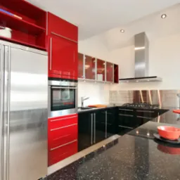 Nowoczesna kuchnia z czerwonymi i czarnymi szafkami na wysoki połysk, stalową lodówką z dystrybutorem wody i czarnym blatem z brokatem, uzupełniona o metalowy backsplash i minimalistyczny okap.