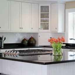 Jasna kuchnia z białymi szafkami, czarnym blatem i wyspą kuchenną z płytą grzewczą, uzupełniona bukietem tulipanów i dekoracyjnymi ceramicznymi kurami.