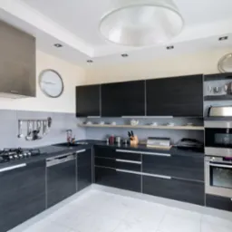 Nowoczesna kuchnia z ciemnymi, drewnopodobnymi szafkami, stalowym okapem i sprzętem AGD, jasnymi płytkami na podłodze i ścianie nad blatem, oświetlona światłem dziennym i lampami sufitowymi.