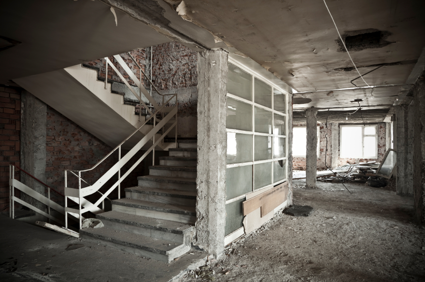 Wnętrze opuszczonego budynku w trakcie remontu: odsłonięta cegła, surowe schody betonowe z białą balustradą, okna z szybami w kratkę, odpadający tynk i gruz na podłodze.