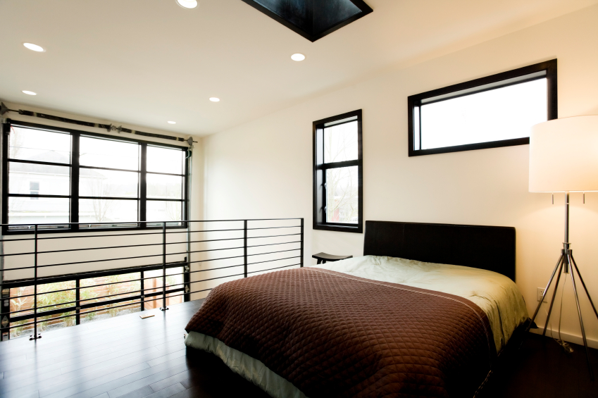 Nowoczesna sypialnia na antresoli z dużym oknem i brązową narzutą w kratę, oświetlona lampą stojącą na trójnogu.