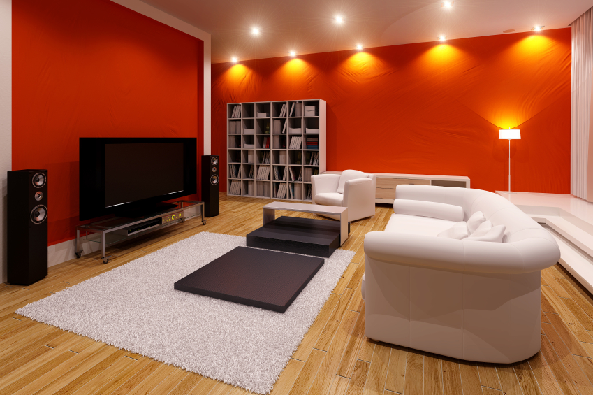 Nowoczesny salon z czerwoną ścianą, białymi meblami, drewnianą podłogą i dużym telewizorem z głośnikami.
