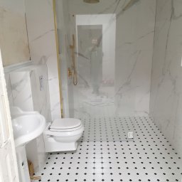 Krzysztof Kozłowski - Wykończona łazienka z białymi płytkami imitującymi marmur, złotą armaturą i czarno-białą mozaiką na podłodze. Kabina prysznicowa ze szklaną ścianką.