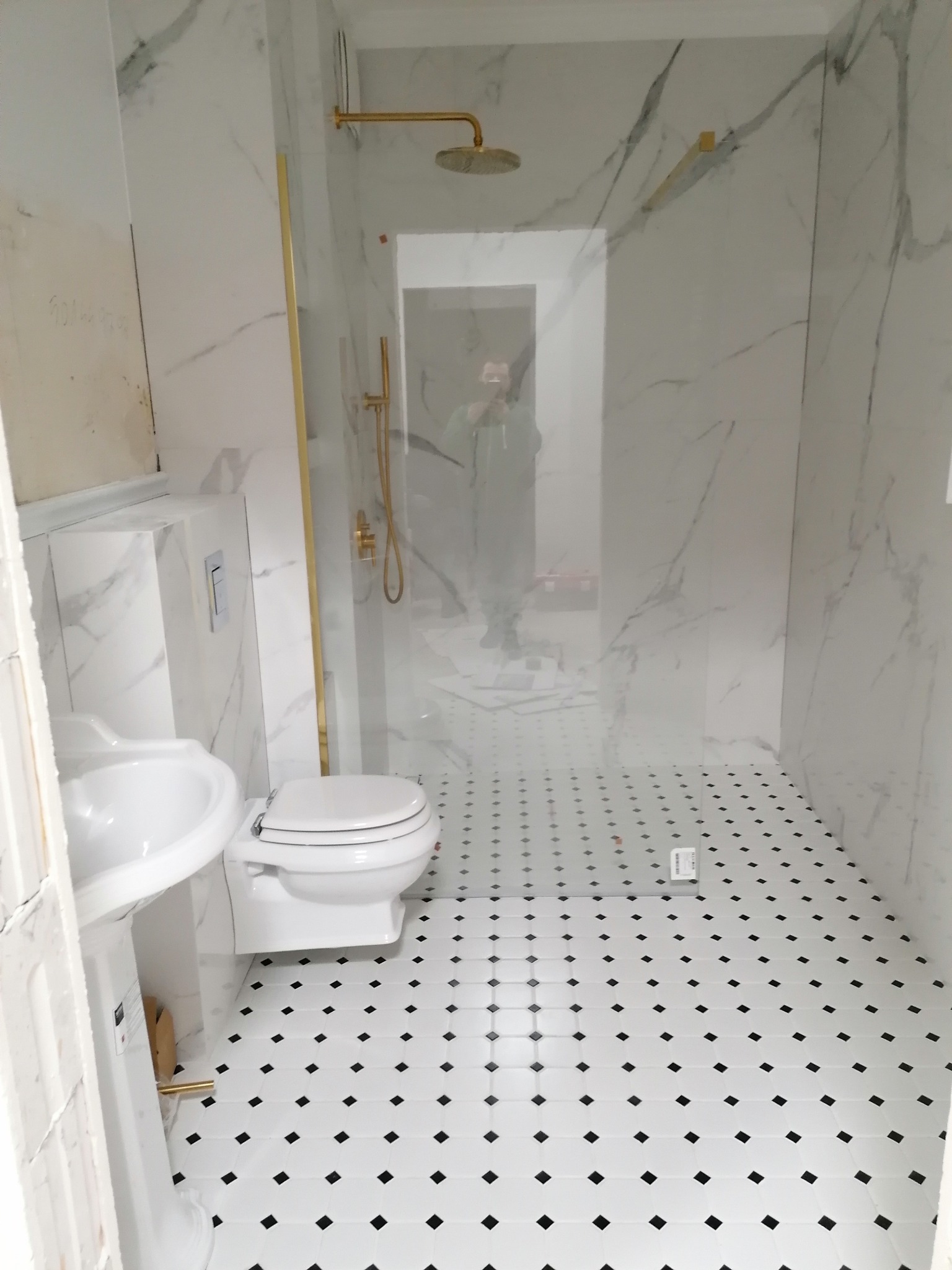 Wykończona łazienka z białymi płytkami imitującymi marmur, złotą armaturą i czarno-białą mozaiką na podłodze. Kabina prysznicowa ze szklaną ścianką.