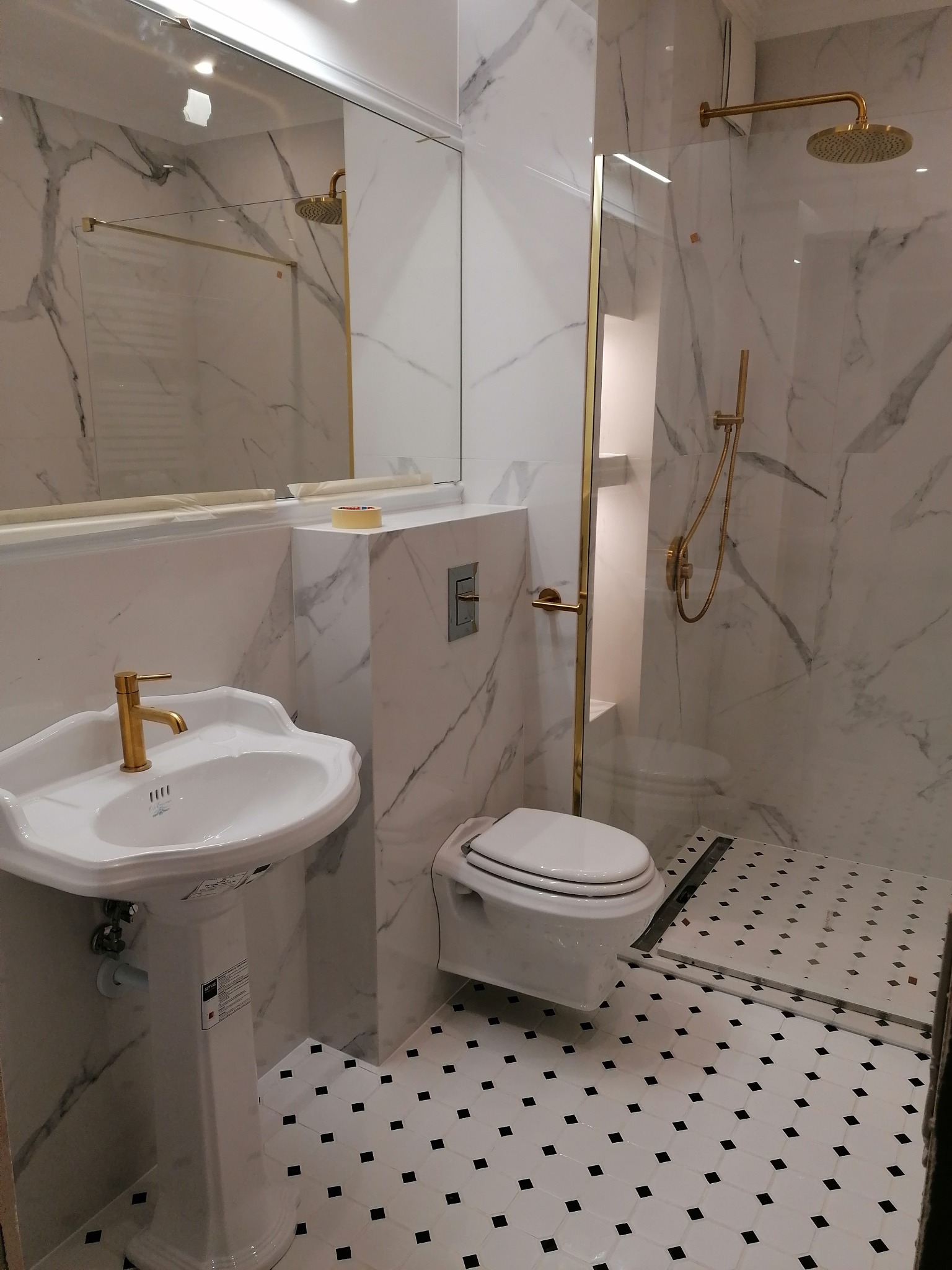 Elegancka łazienka z marmurowymi ścianami, złotą armaturą i czarno-białą mozaiką podłogową. Umywalka na postumencie, toaleta podwieszana i kabina prysznicowa.