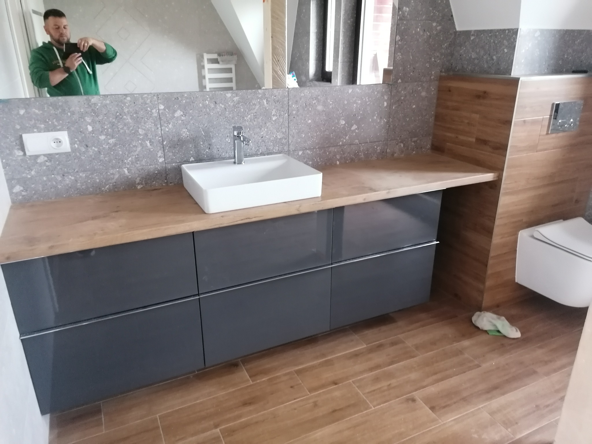 Nowoczesna łazienka z szarą zabudową pod umywalką, drewnianym blatem i płytkami imitującymi drewno. W lustrze odbija się osoba robiąca zdjęcie.