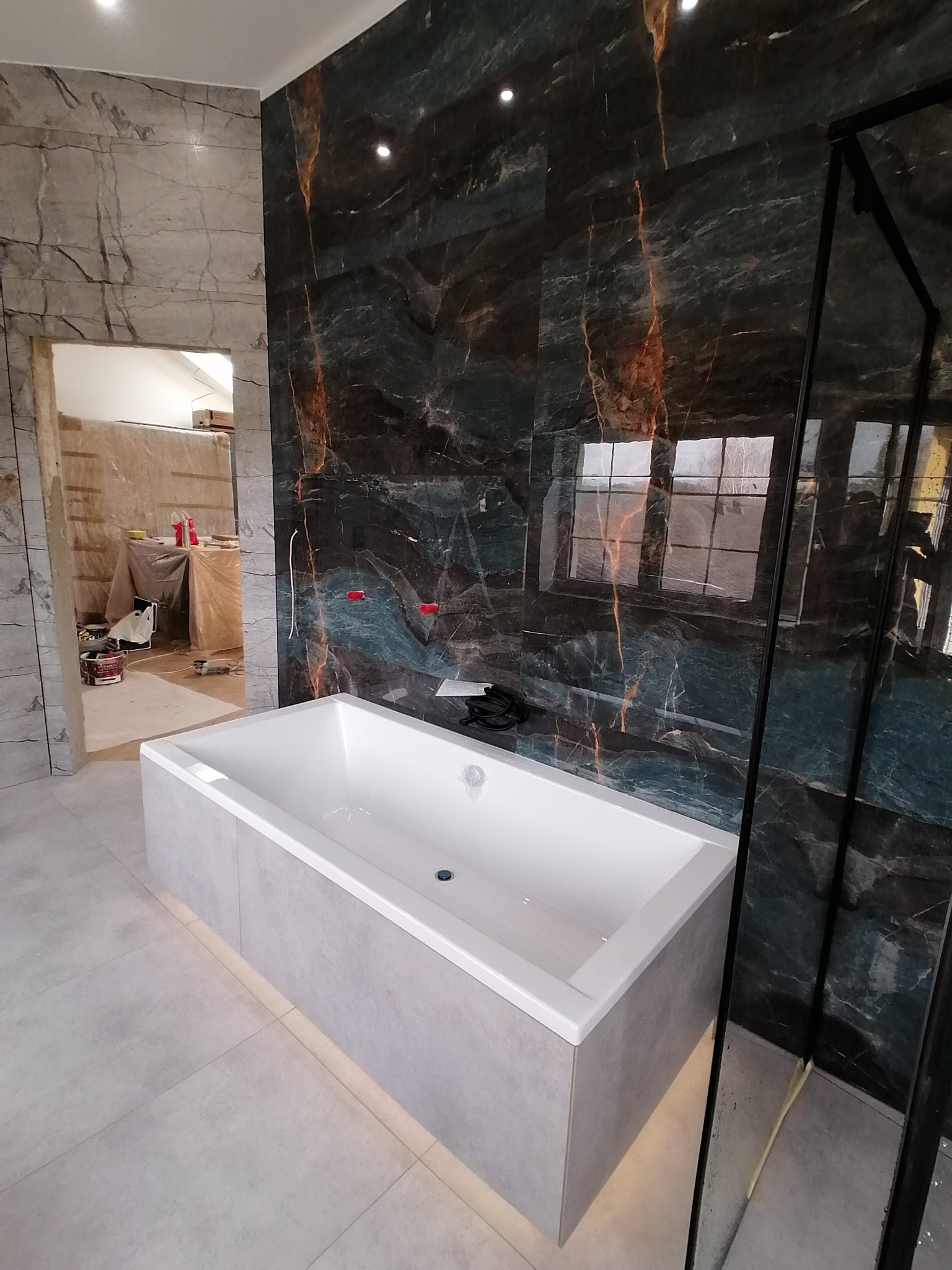 Nowoczesna łazienka z wanną w zabudowie, wykończona płytkami imitującymi marmur w odcieniach szarości i zieleni. Widoczna kabina prysznicowa i wejście do innego pomieszczenia.