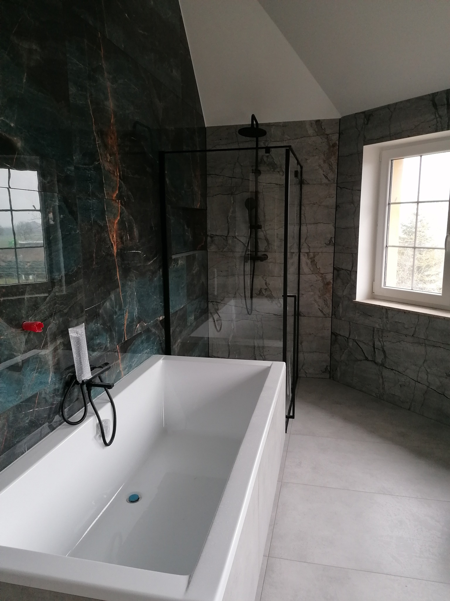 Nowoczesna łazienka z wanną, kabiną prysznicową typu walk-in z czarnymi profilami i marmurowymi płytkami w odcieniach szarości i zieleni. Okno z widokiem na zewnątrz.