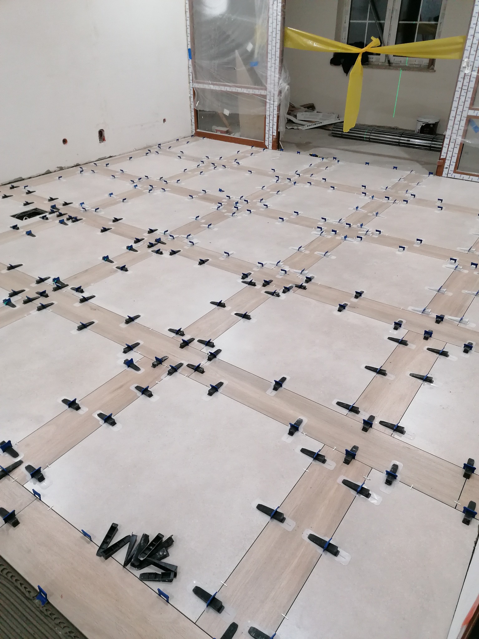 Układanie jasnych, kwadratowych płytek podłogowych z użyciem niebieskich klipsów poziomujących. Widoczne drzwi balkonowe zabezpieczone folią.