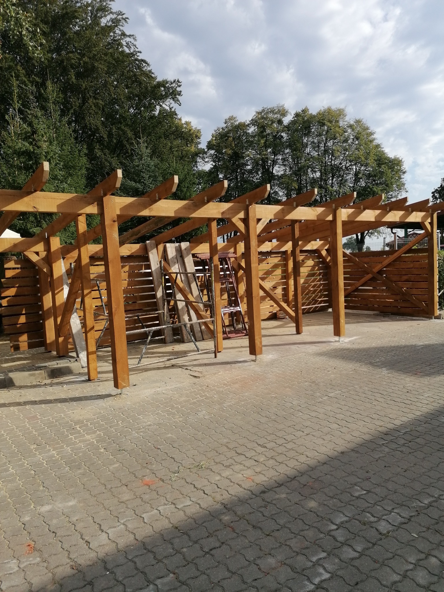 Drewniana konstrukcja zadaszenia tarasu w Bartoszycach, widok z zewnątrz. Elementy konstrukcyjne z belek, w tle drzewa i ogrodzenie. Widoczne drabiny i rusztowania.