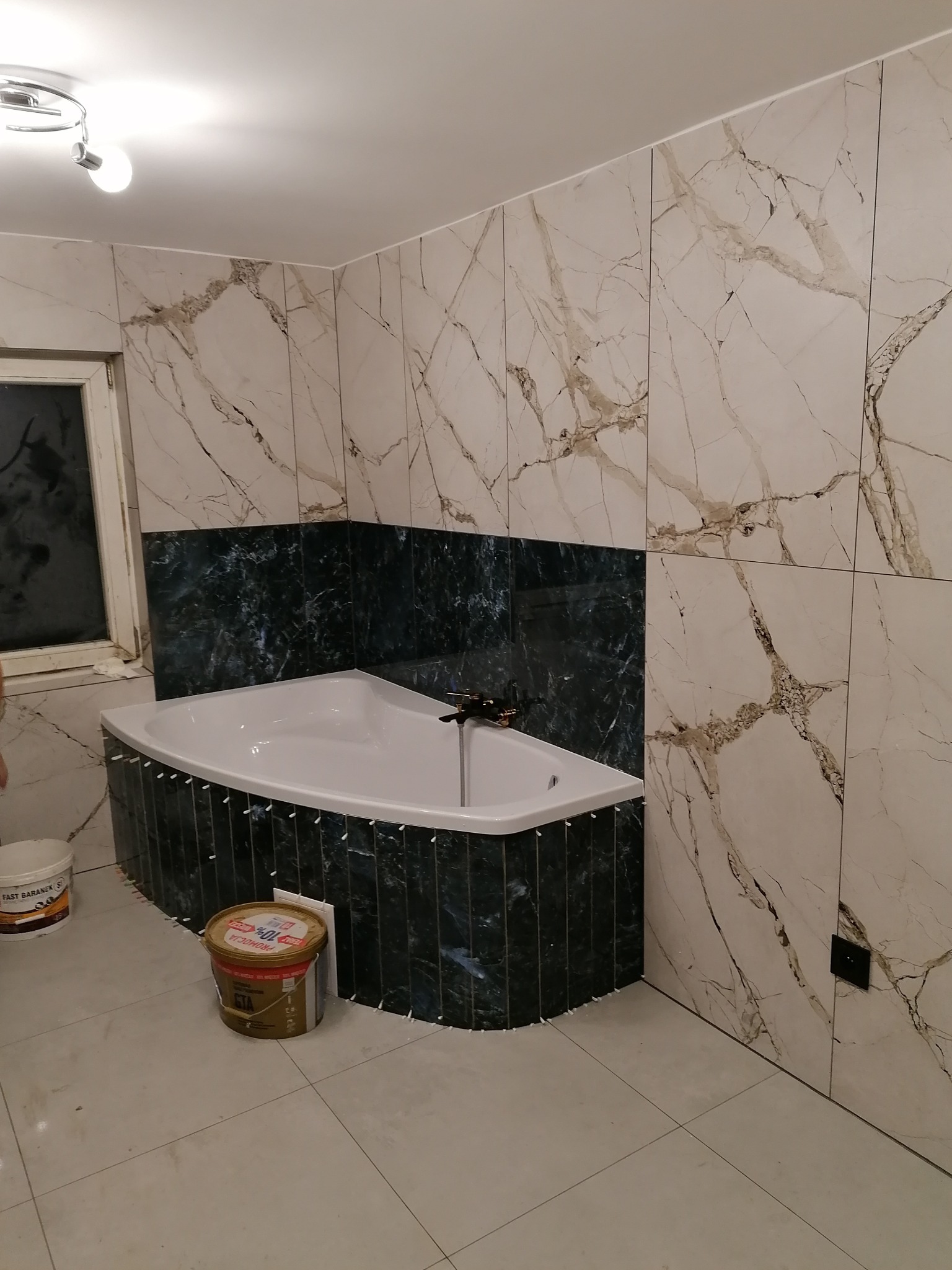 Wnętrze łazienki w trakcie wykańczania: biała wanna obłożona ciemnozielonymi płytkami, ściany wyłożone płytkami imitującymi marmur, widoczne narzędzia glazurnicze.