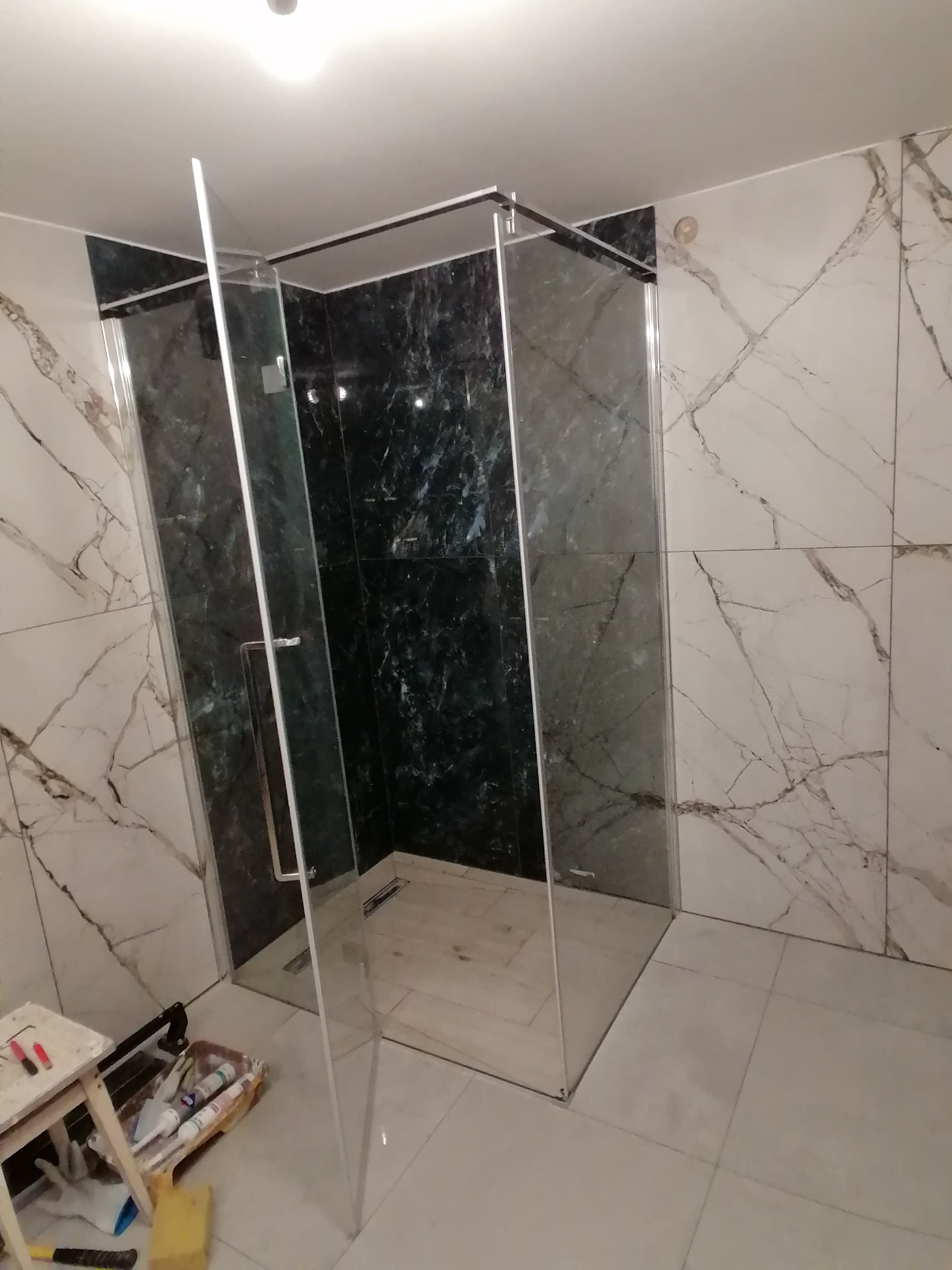 Nowoczesna kabina prysznicowa z transparentnego szkła, wykończona czarnym marmurem i białymi płytkami imitującymi marmur. Widoczne narzędzia glazurnicze.