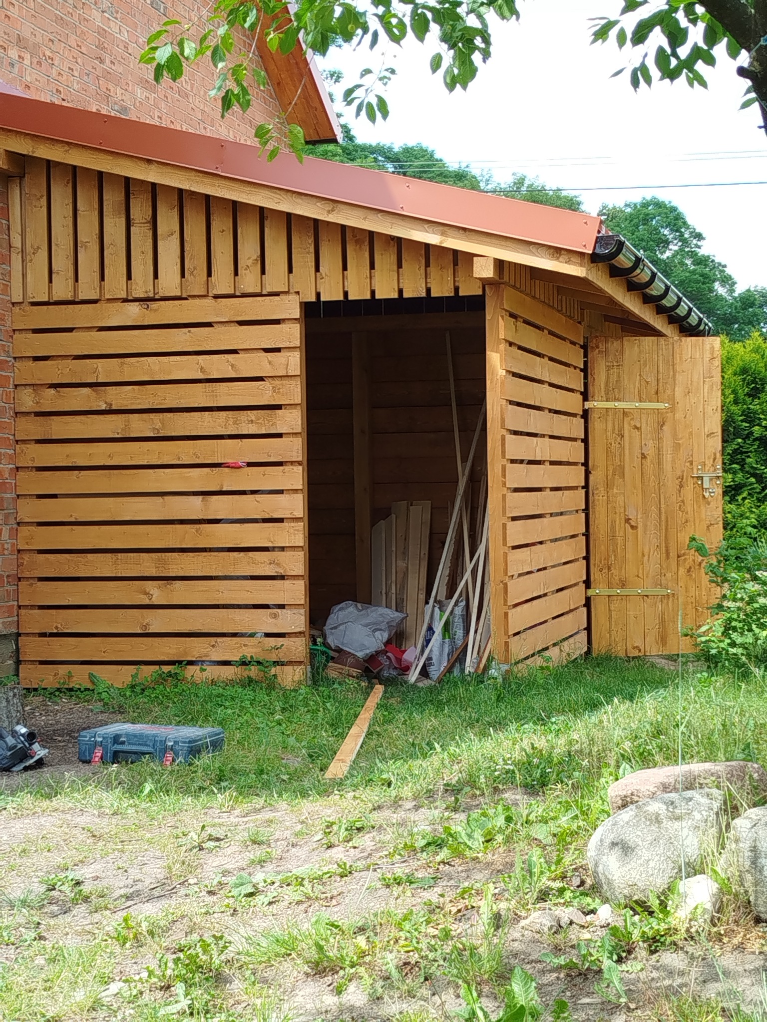 Drewniana konstrukcja z otwartymi drzwiami, widoczne narzędzia i materiały budowlane wewnątrz. Konstrukcja przylega do ceglanego budynku, otoczona zielenią.