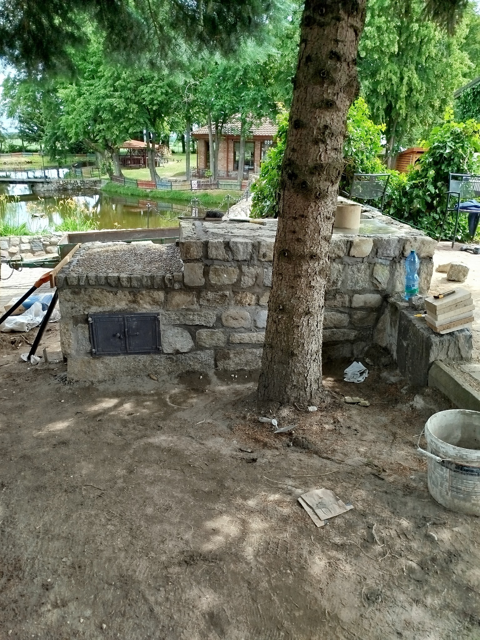 Kamienny grill ogrodowy w trakcie budowy, z metalowymi drzwiczkami i drzewem wrastającym w konstrukcję, na tle stawu i zieleni.