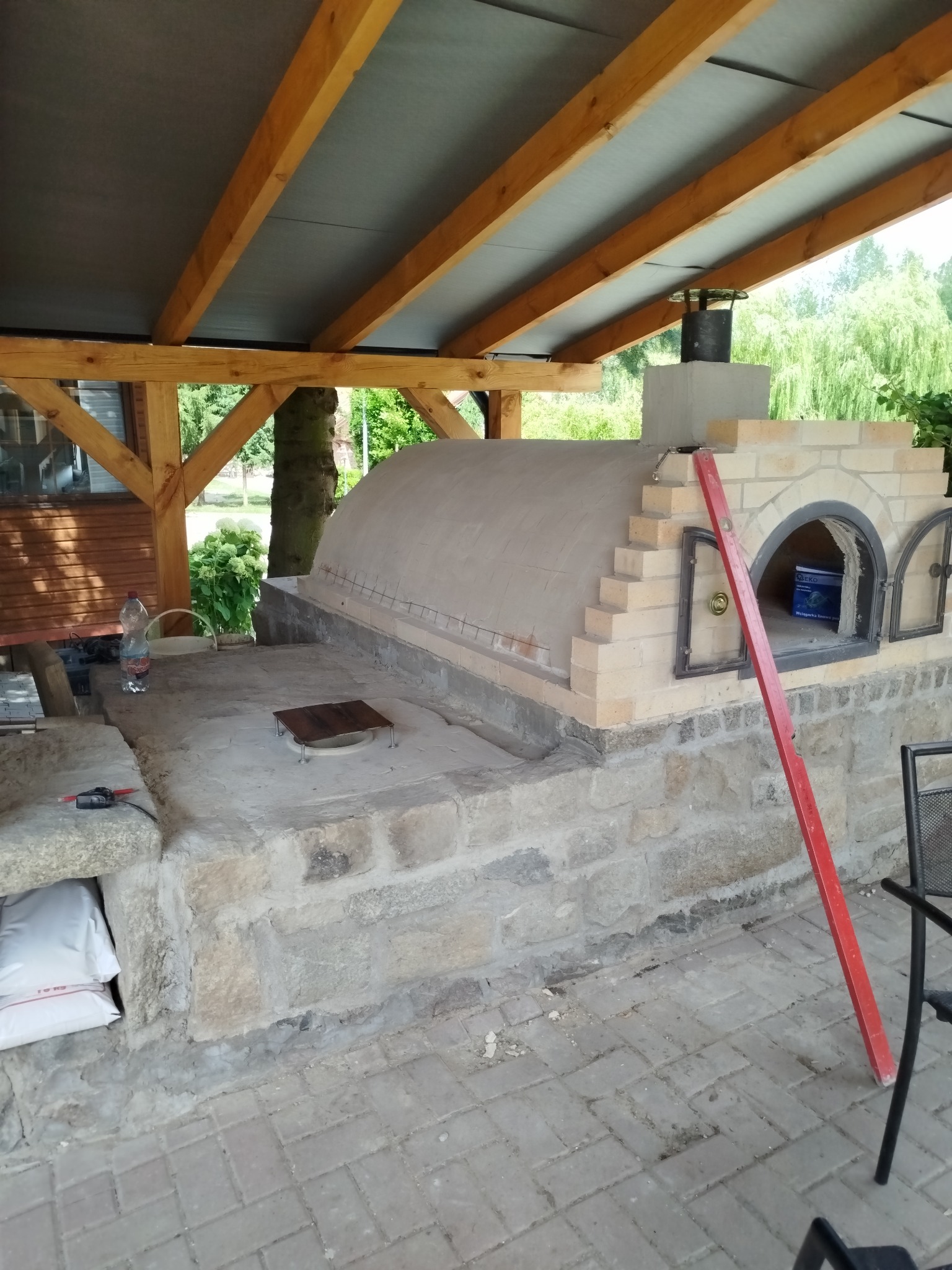 Zewnętrzny piec chlebowy z cegły pod drewnianą konstrukcją zadaszenia, z widocznymi narzędziami i materiałami budowlanymi w otoczeniu ogrodu.