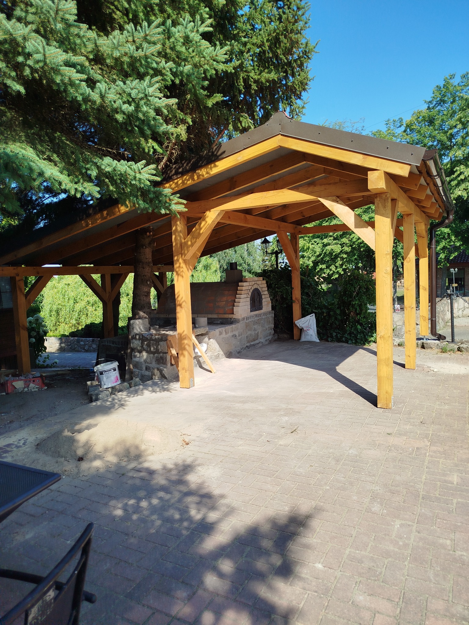 Drewniana altana z piecem chlebowym w ogrodzie, konstrukcja zadaszona, ceglany piec, drzewo wkomponowane w konstrukcję, kostka brukowa na ziemi.