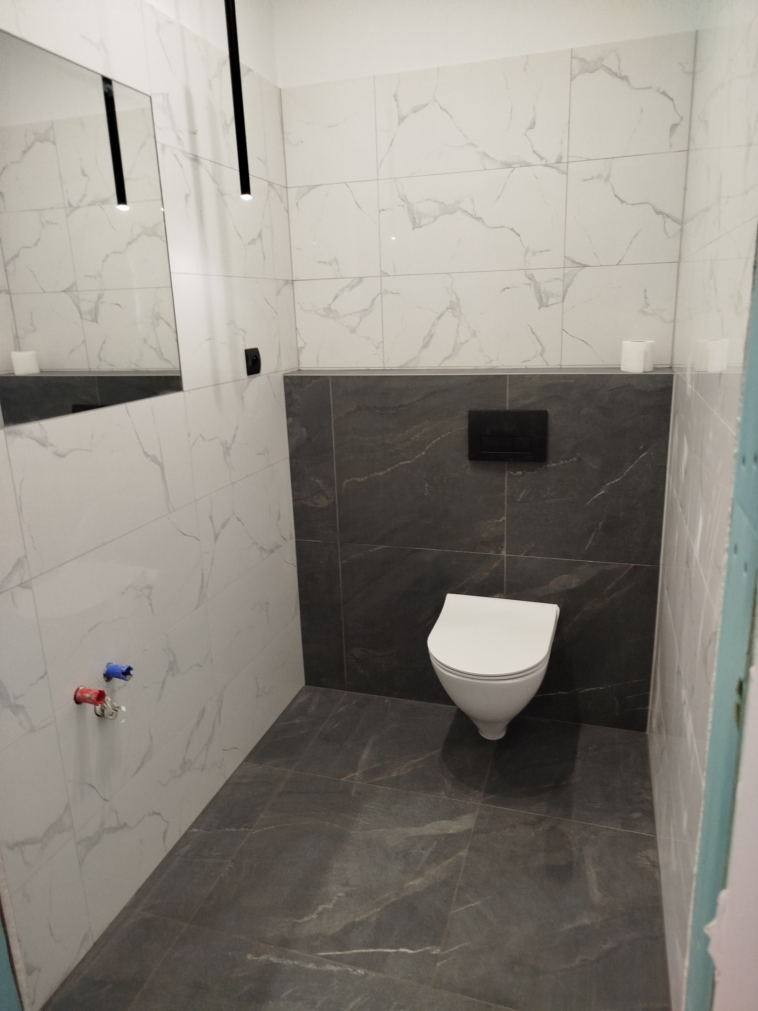 Nowoczesna toaleta z białymi i szarymi płytkami imitującymi marmur, wisząca muszla klozetowa i minimalistyczne oświetlenie.