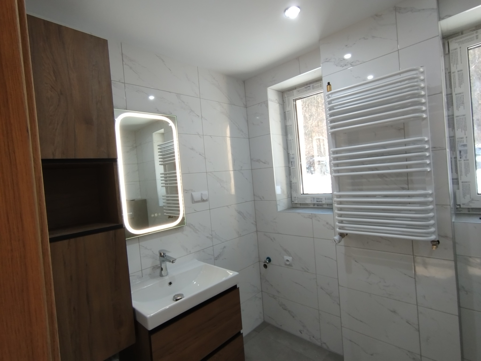 Nowoczesna łazienka z białymi marmurkowymi płytkami, drewnianą szafką pod umywalką i podświetlanym lustrem. Grzejnik drabinkowy obok okna.