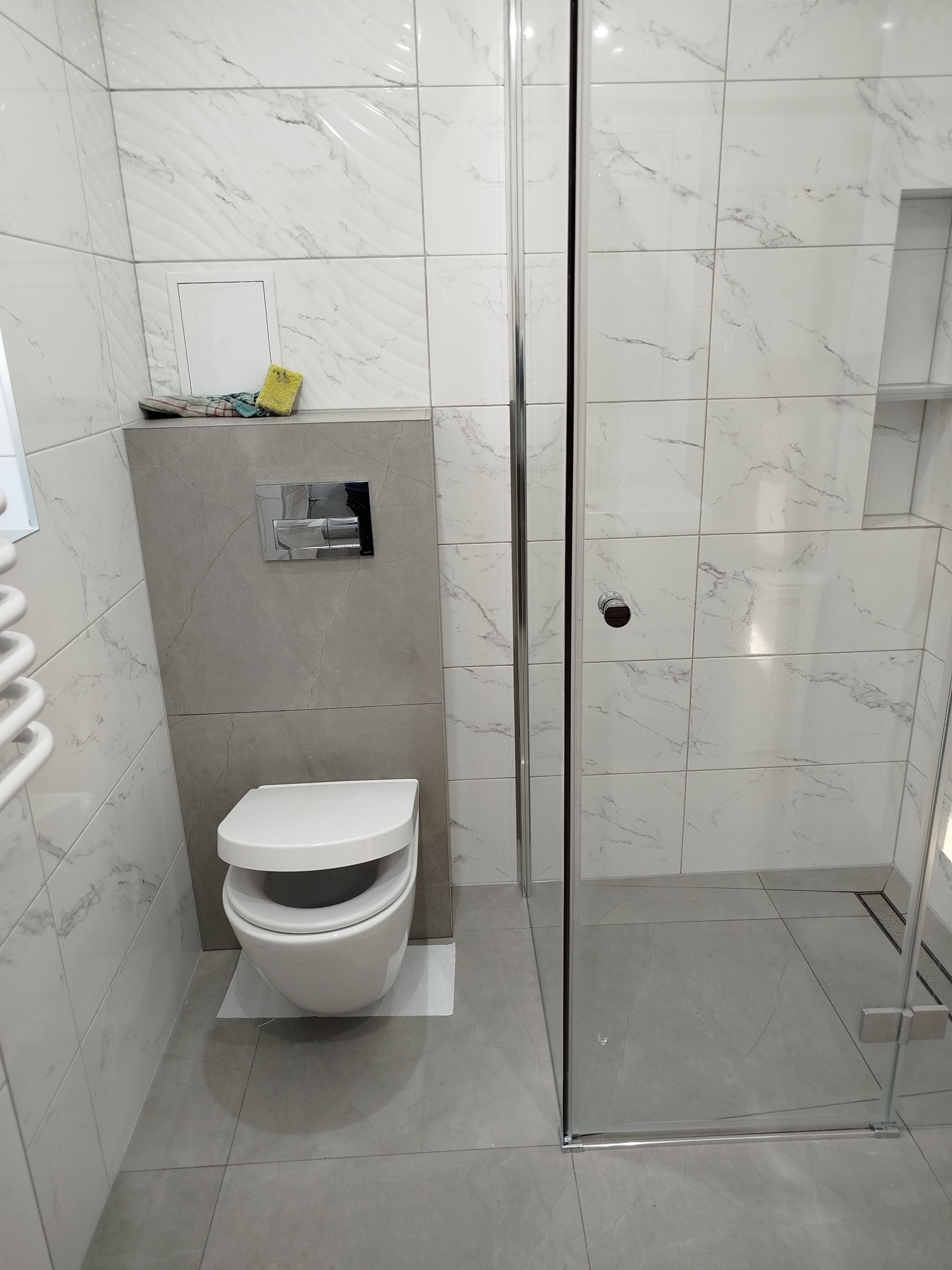 Nowoczesna łazienka z kabiną prysznicową typu walk-in, białą ceramiką i szarymi płytkami imitującymi marmur. Widoczny fragment grzejnika i akcesoria łazienkowe.