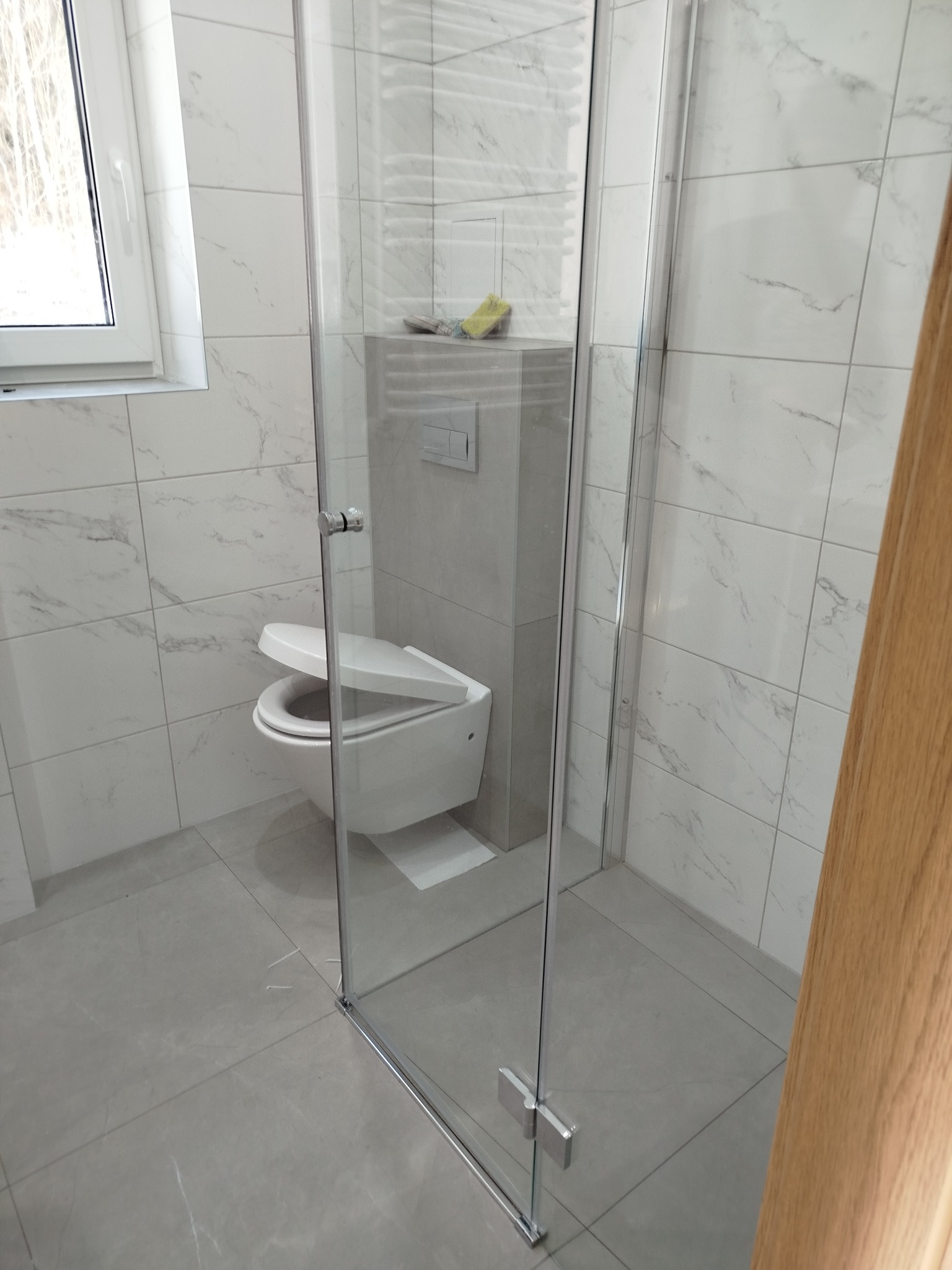 Nowoczesna łazienka po remoncie w Bartoszycach. Kabina prysznicowa ze szkła, białe płytki imitujące marmur, podwieszana toaleta. Minimalistyczny design i funkcjonalność.