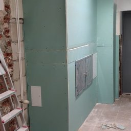 PRZEDSIĘBIORSTWO BUDOWLANE NATKA SPÓŁKA Z OGRANICZONĄ ODPOWIEDZIALNOŚCIĄ - Montaż płyt karton-gips na ścianie z widocznymi otworami rewizyjnymi. W tle surowa cegła i drabina. Podłoga częściowo zakryta gruzem.