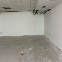 PRZEDSIĘBIORSTWO BUDOWLANE NATKA SPÓŁKA Z OGRANICZONĄ ODPOWIEDZIALNOŚCIĄ - Wnętrze w trakcie remontu: białe ściany, surowa podłoga z resztkami zaprawy, odsłonięta instalacja sufitowa. Butelka na podłodze dodaje realizmu.