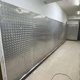 PRZEDSIĘBIORSTWO BUDOWLANE NATKA SPÓŁKA Z OGRANICZONĄ ODPOWIEDZIALNOŚCIĄ - Korytarz z wyłożonymi ścianami blachą ryflowaną, zabezpieczającą przed uszkodzeniami. Widoczne drzwi ewakuacyjne i elementy wykończeniowe.