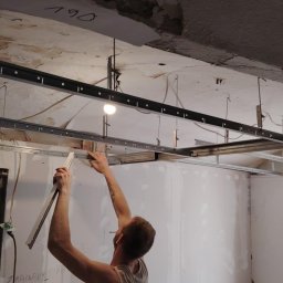 PRZEDSIĘBIORSTWO BUDOWLANE NATKA SPÓŁKA Z OGRANICZONĄ ODPOWIEDZIALNOŚCIĄ - Montaż metalowego stelażu sufitu podwieszanego w pomieszczeniu z oknami. Widoczne profile, ręce montera i fragment ściany z instalacją elektryczną.