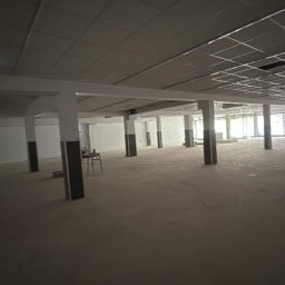 PRZEDSIĘBIORSTWO BUDOWLANE NATKA SPÓŁKA Z OGRANICZONĄ ODPOWIEDZIALNOŚCIĄ - Przestronne wnętrze w trakcie prac wykończeniowych: surowy beton na podłodze, widoczne kolumny i instalacje pod sufitem. Osoba w oddali dodaje skali.