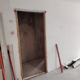 PRZEDSIĘBIORSTWO BUDOWLANE NATKA SPÓŁKA Z OGRANICZONĄ ODPOWIEDZIALNOŚCIĄ - Przygotowany otwór drzwiowy z ościeżnicą, wypełniony pianką montażową, poziomica oparta o ścianę, widoczne narzędzia i materiały budowlane. Surowy stan wykończenia.