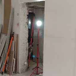 PRZEDSIĘBIORSTWO BUDOWLANE NATKA SPÓŁKA Z OGRANICZONĄ ODPOWIEDZIALNOŚCIĄ - Wnętrze w trakcie remontu: oświetlona lampami ściana po szpachlowaniu, widoczne płyty karton-gips i worki z zaprawą. Przejście w stanie surowym.
