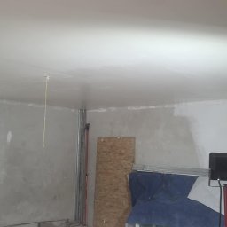 PRZEDSIĘBIORSTWO BUDOWLANE NATKA SPÓŁKA Z OGRANICZONĄ ODPOWIEDZIALNOŚCIĄ - Wnętrze w trakcie szpachlowania: gładki sufit, częściowo wykończone ściany, okno z widokiem na zieleń, narzędzia malarskie i lampa budowlana.