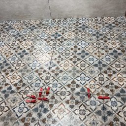 PRZEDSIĘBIORSTWO BUDOWLANE NATKA SPÓŁKA Z OGRANICZONĄ ODPOWIEDZIALNOŚCIĄ - Ułożona podłoga z płytek w kwiatowy wzór, z czerwonymi klipsami poziomującymi. Widoczne fragmenty ściany z brązowymi i szarymi płytkami.