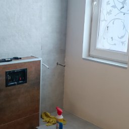 Wykończona łazienka z szarymi płytkami, widoczne narzędzia i środki czystości. Umywalka w zabudowie z brązowymi elementami. Okno z dekoracyjną folią.