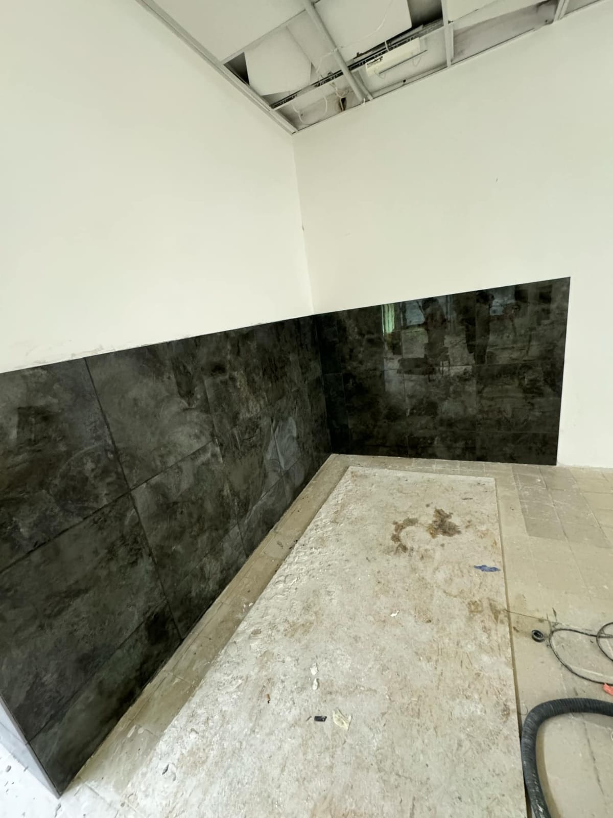 Częściowo wykończona ściana z ciemnych, błyszczących płytek w pomieszczeniu z surowym sufitem i przygotowanym podłożem pod posadzkę. Widoczne narzędzia.