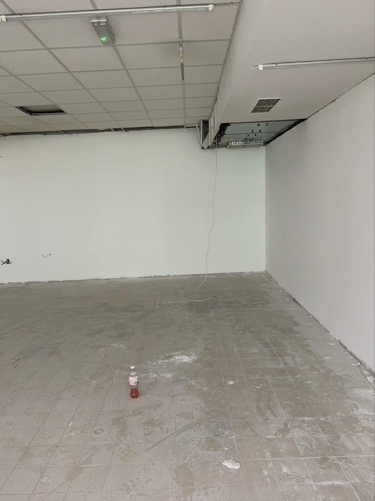 Wnętrze w trakcie remontu: białe ściany, surowa podłoga z resztkami zaprawy, odsłonięta instalacja sufitowa. Butelka na podłodze dodaje realizmu.