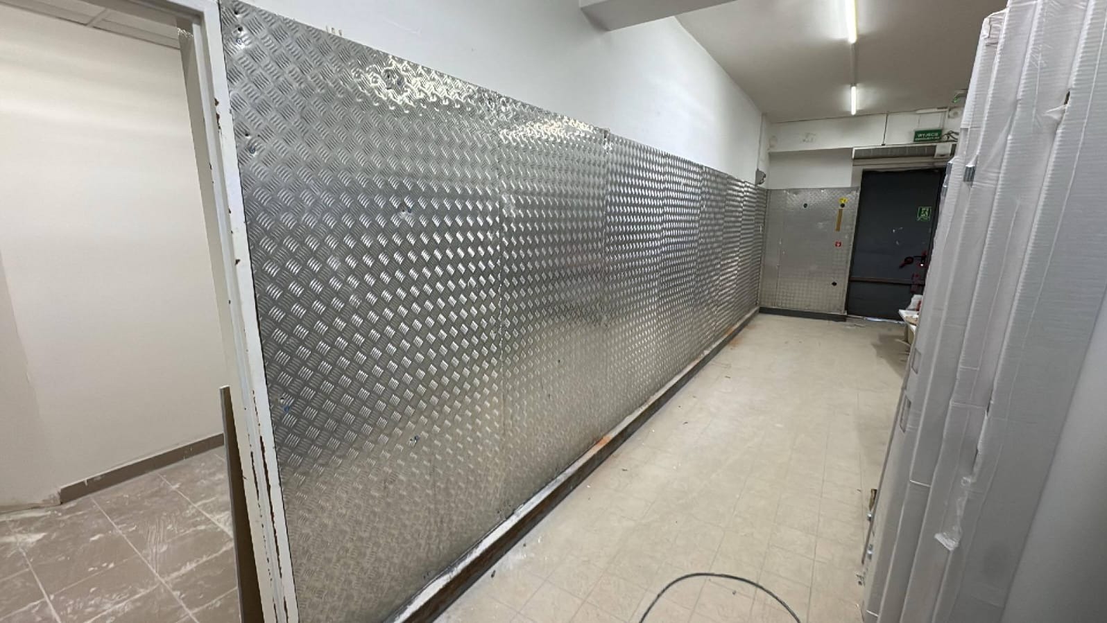 Korytarz z wyłożonymi ścianami blachą ryflowaną, zabezpieczającą przed uszkodzeniami. Widoczne drzwi ewakuacyjne i elementy wykończeniowe.