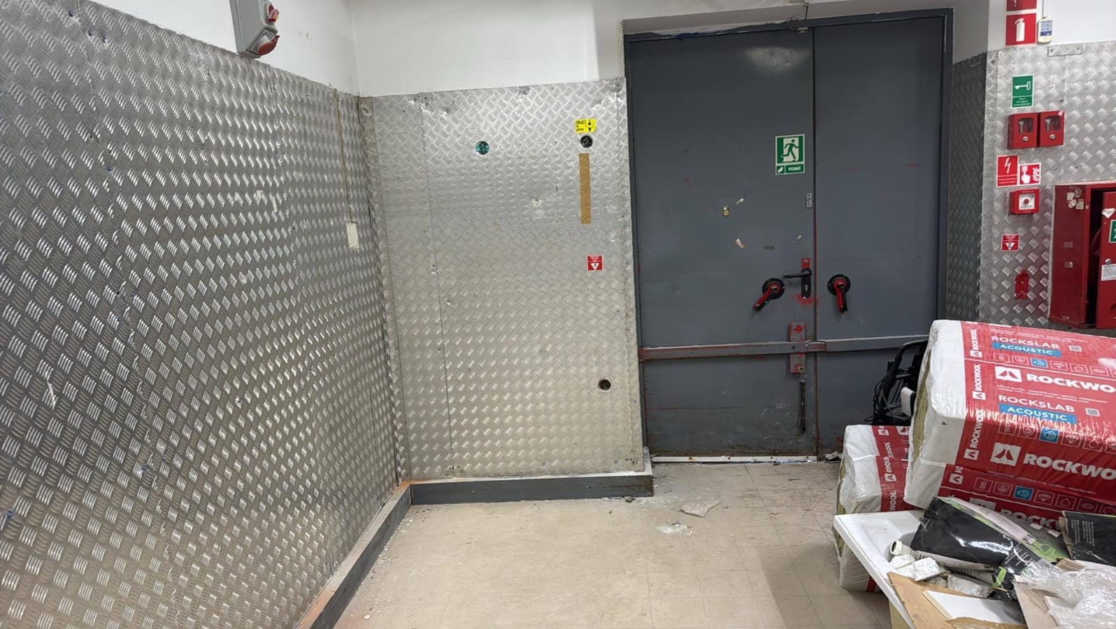 Wnętrze techniczne z blachą ryflowaną na ścianach, stalowymi drzwiami ewakuacyjnymi i materiałami izolacyjnymi Rockwool. Widoczne oznaczenia bezpieczeństwa i instalacje.