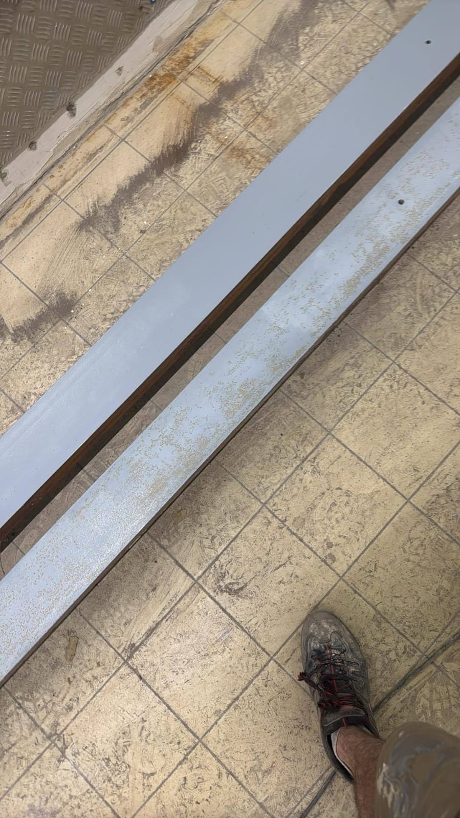 Dwie metalowe belki pomalowane szarą farbą, z widocznymi śladami korozji i odpryskami farby, leżące na podłodze wyłożonej kwadratowymi płytkami. Widoczny fragment buta.