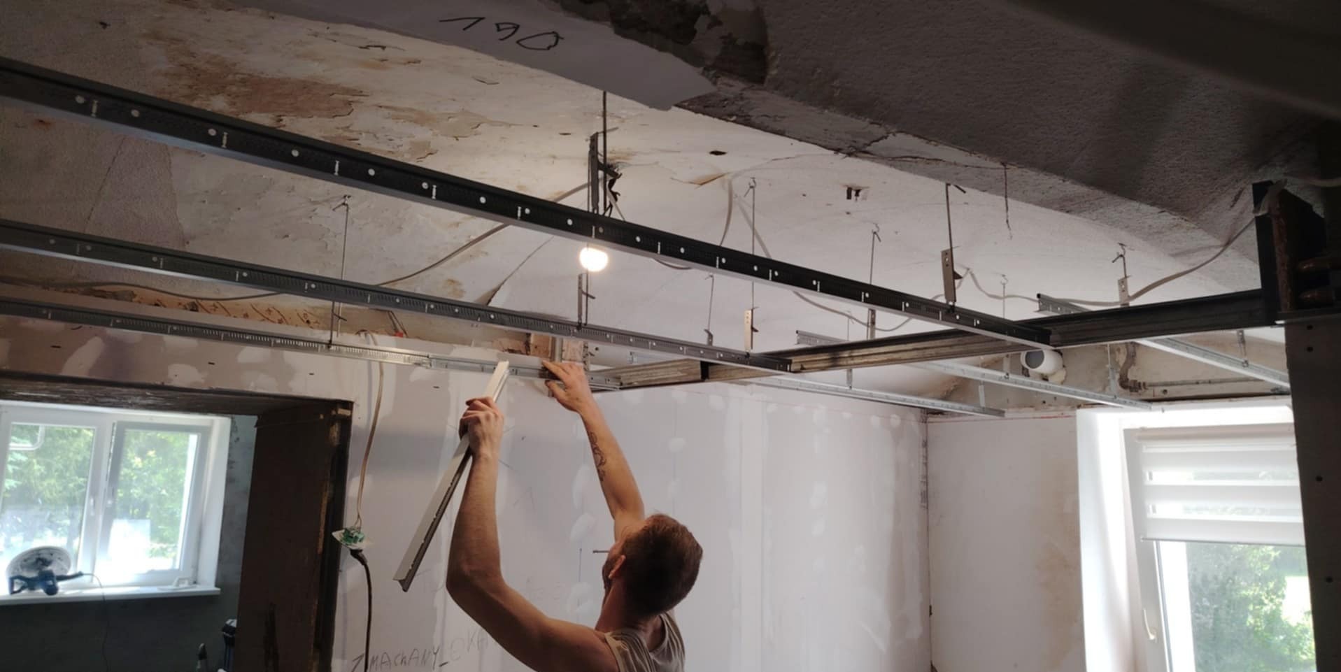 Montaż metalowego stelażu sufitu podwieszanego w pomieszczeniu z oknami. Widoczne profile, ręce montera i fragment ściany z instalacją elektryczną.