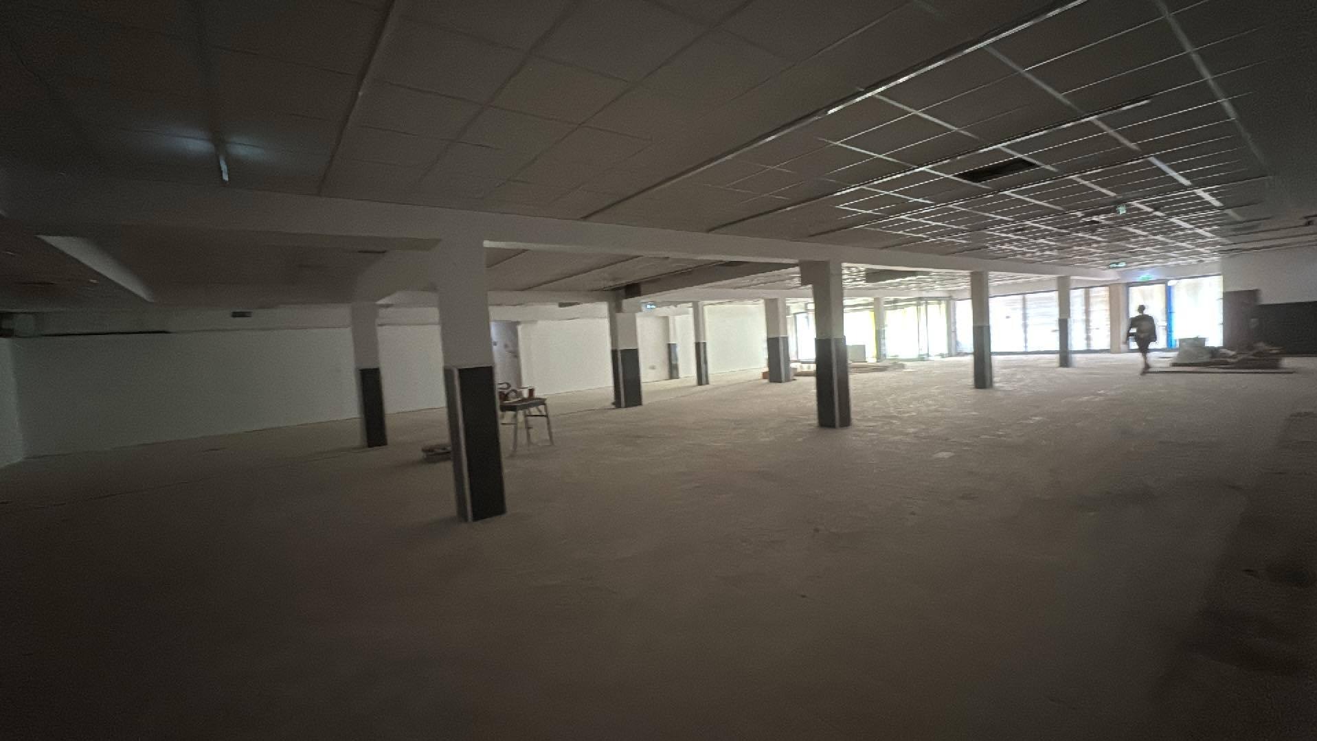 Przestronne wnętrze w trakcie prac wykończeniowych: surowy beton na podłodze, widoczne kolumny i instalacje pod sufitem. Osoba w oddali dodaje skali.